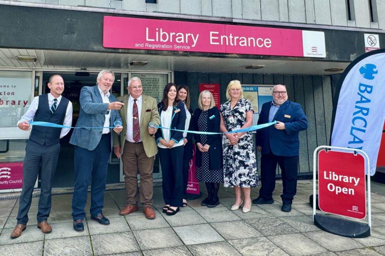 A group of men and women in business attire cut a ribbon outside a library.