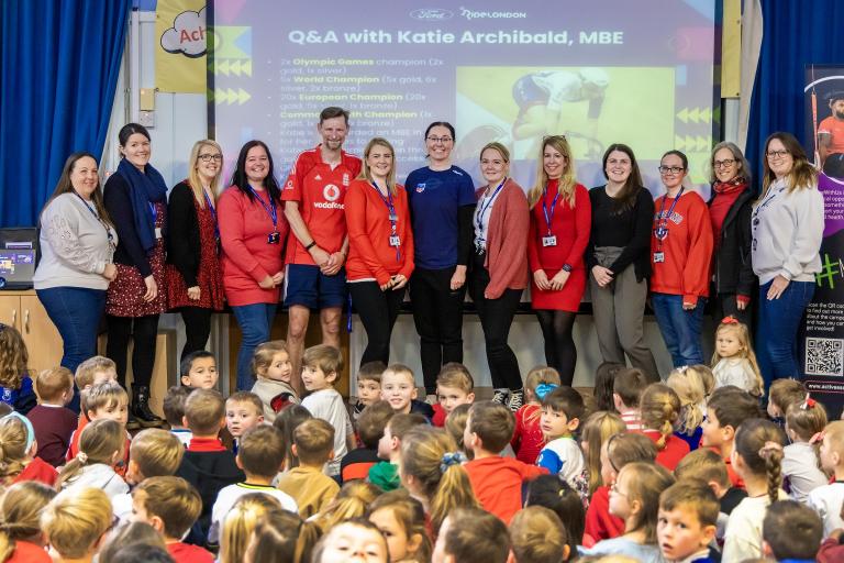 A row of people standing in front of a projector screen facing the camera. They are standing in front of a large group of schoolchildren who are sitting on the ground.